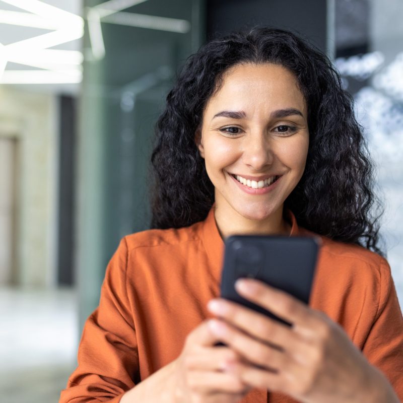 Beautiful woman in the office, happy and smiling latin american business woman uses internet phone close up, female worker reads message and browses internet pages inside office building.