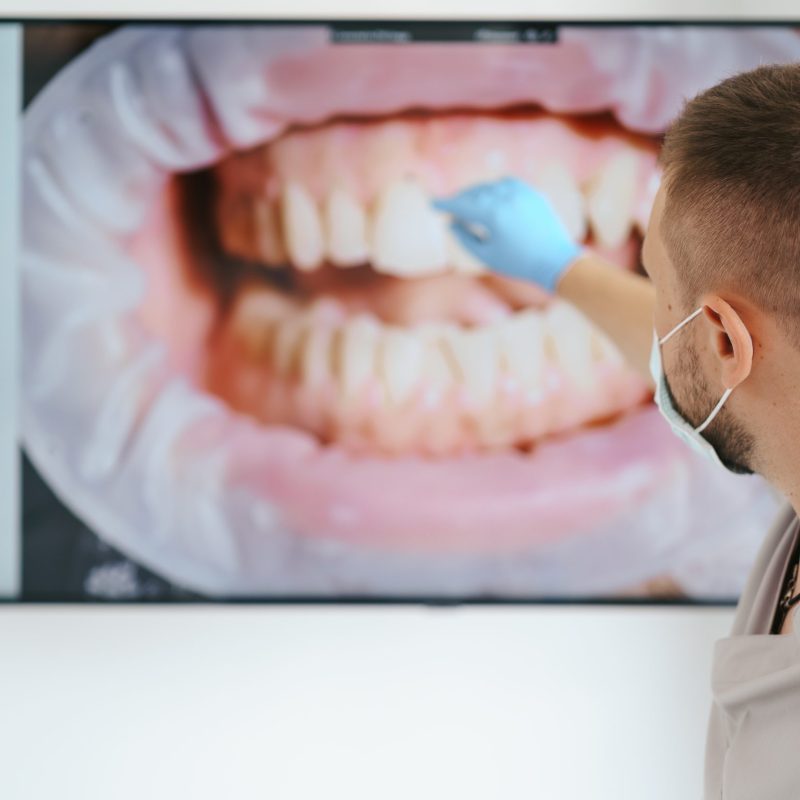 Dentist in medical mask pointing on the patient teeth on a big screen explaining treatment phases