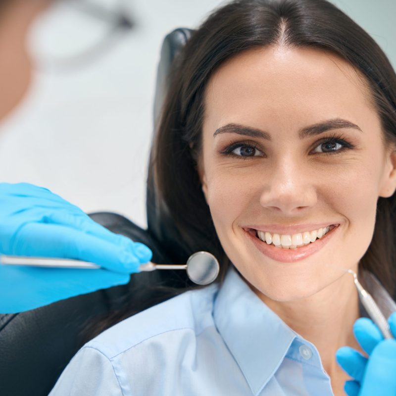 Smiling woman patient sitting in orthodontic chair and waiting qualified dentist examining her teeth and gums with special mouth mirror and sickle probe